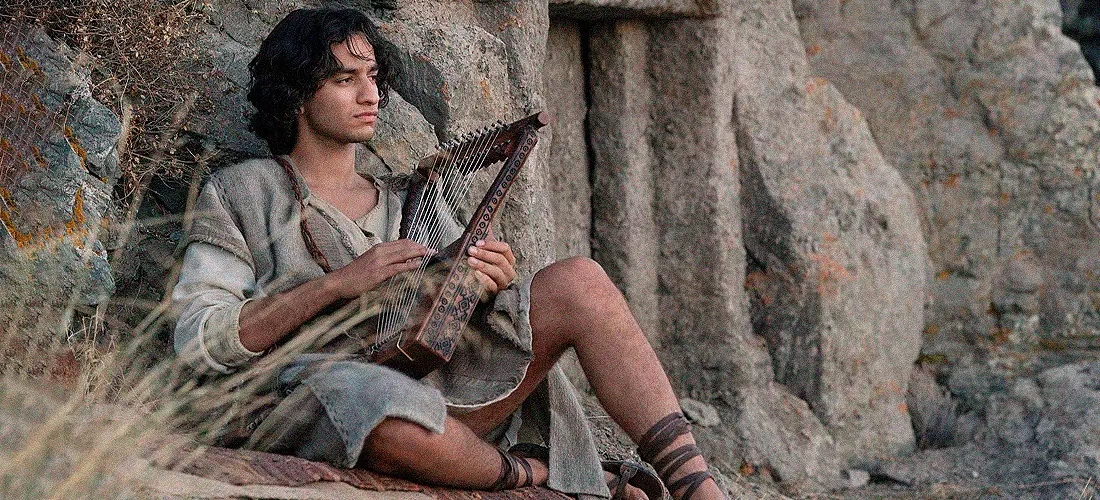 Кадр из сериала «House of David»: юноша в древней одежде играет на арфе, сидя у скального храма, окружённый природным ландшафтом.